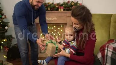 母亲、父亲和小婴儿坐在装饰圣诞树附近。 爸爸把盒子送给坐在妈妈旁边的孩子